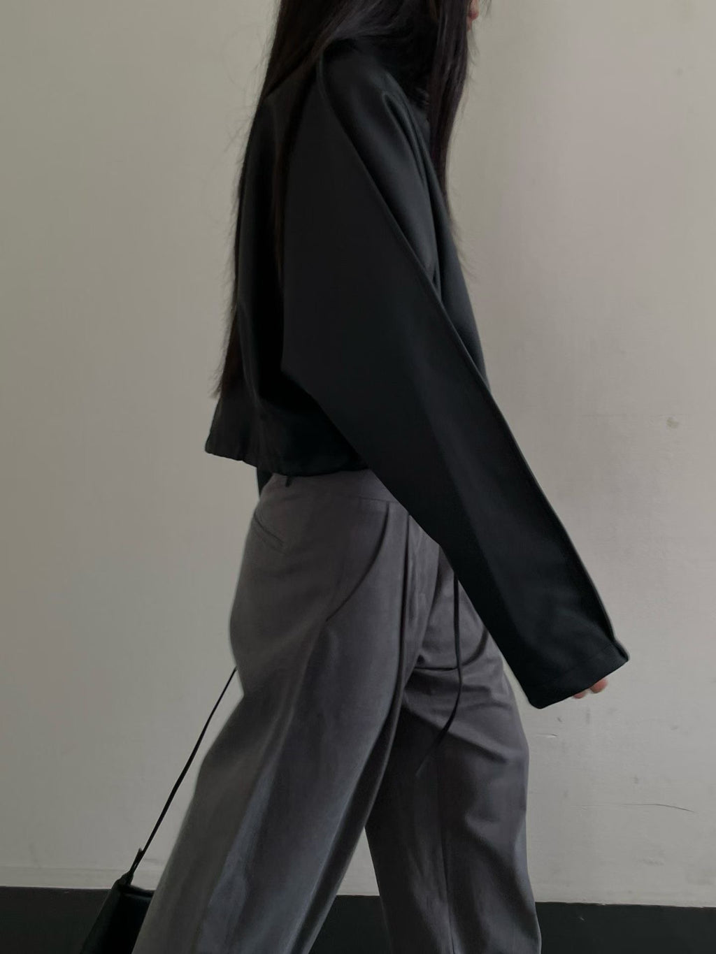 Side view of black cropped faux leather jacket with long sleeves, styled with grey trousers