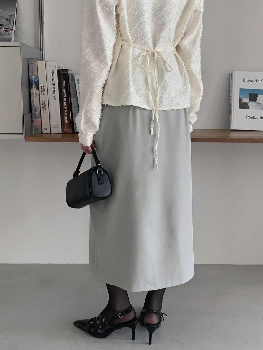 Rear view of a beige midi skirt styled with an ivory tie-back top and black heels.