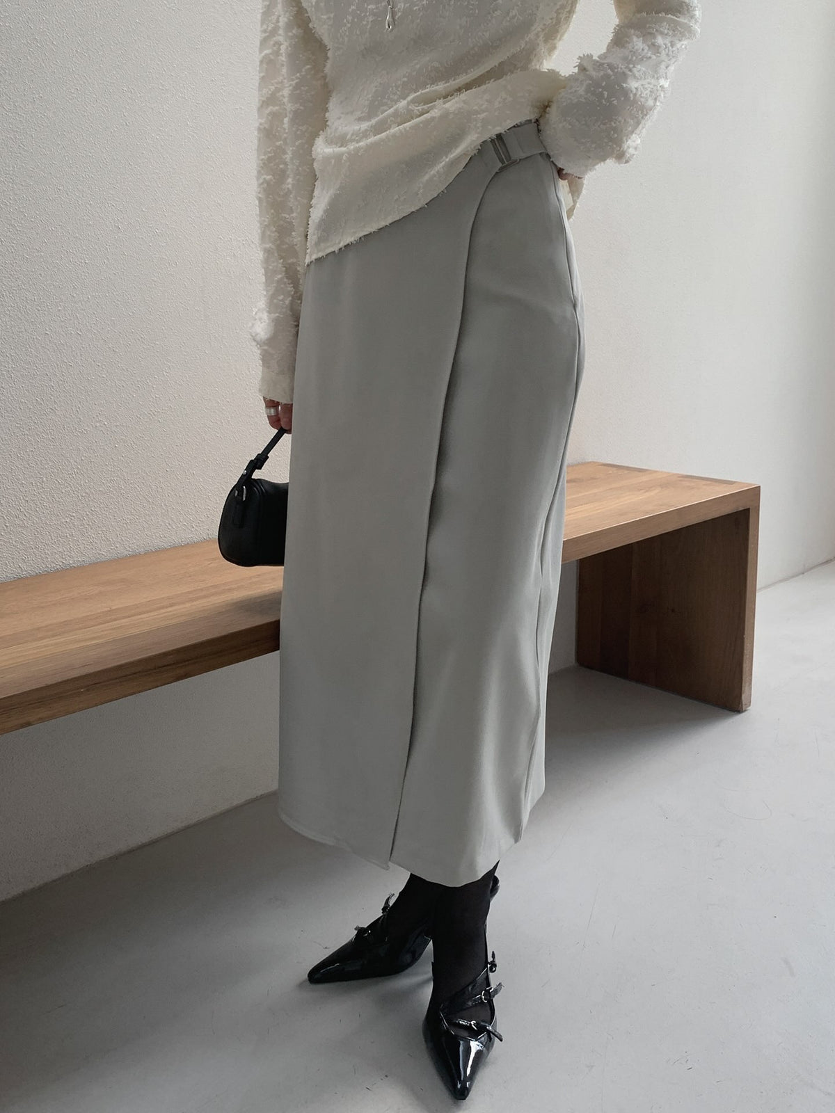 Woman wearing a beige buckle-detail midi skirt with an ivory top and black heels beside a wooden bench.
