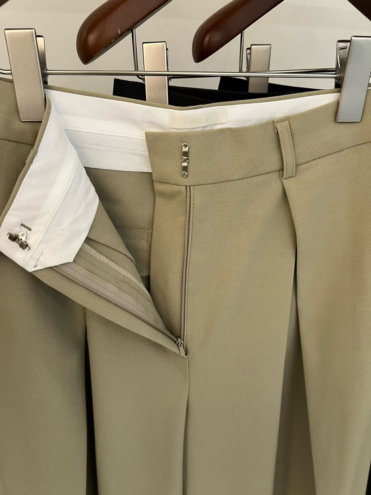 Close-up of beige pants showing the open zip fly, hook fastening and waistband interior.